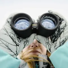 Kobieta w rękawiczkach z wzorem gałęzi patrzy przez lornetkę Nikon, podziwiając górski krajobraz. Idealna lornetka w góry.