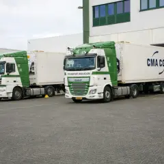 Zwei weiße und grüne LKWs stehen bereit, um entladen zu werden. Einer trägt die Aufschrift "Transport Rien Boom", der andere "CMA CGM".