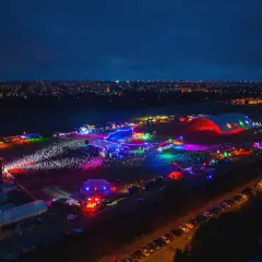 Nocne, kolorowe światła rozświetlają teren festiwalu na powietrzu. Tłumy bawią się przy scenach i namiotach, a w tle widać miasto.