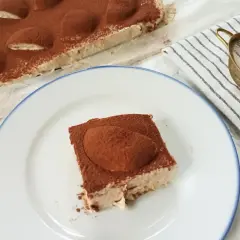 Pyszne ciasto cappuccino z mascarpone, posypane kakao, czeka na talerzu. Obok całe ciasto i sitko do posypywania.