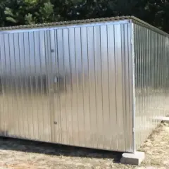 Nowy garaż blaszany z dwuskrzydłowymi drzwiami, stojący na betonowych bloczkach, na tle drzew.