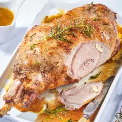 Gigot d'agneau désossé au four, doré et juteux, garni de romarin et d'ail. Servi avec sa sauce dans un plat blanc.