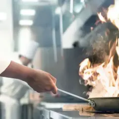Ein Koch wirbelt eine brennende Pfanne in einer professionellen Küche. Flammen schlagen hoch und erzeugen viel Rauch.