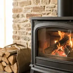 Ein gemütlicher Dauerbrandofen mit brennendem Holz vor einer Steinwand. Daneben liegt ein Stapel Holzscheite in einem Tragekorb.