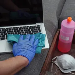 Ręka w niebieskiej rękawiczce czyści laptopa. Obok butelka z napisem "OBJECT" i maseczka. Denaturat jest do dezynfekcji przedmiotów.