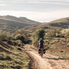 Ciclista pedaleando por un camino rural en España, disfrutando de sus viajes en bicicleta por España. Paisaje montañoso y vegetación verde.