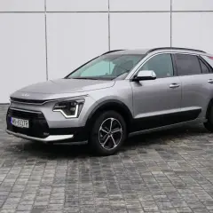 Srebrny Kia Niro na tle białej ściany. Wiele osób szuka kia niro opinie przed zakupem.