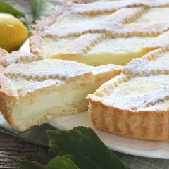 Una fetta di crostata di ricotta, cosparsa di zucchero a velo, accanto a un limone e foglie verdi su un tavolo di legno.