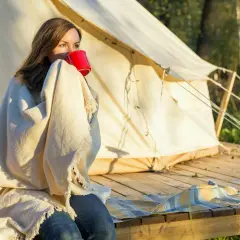 Kvinna med filt och röd mugg vid tältet. En mysig morgon som visar vikten av en bra packlista camping.