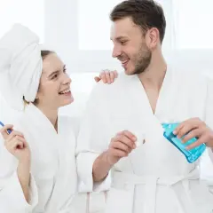 Coppia sorridente in accappatoio, lei con spazzolino, lui versa collutorio. Imparare come usare il collutorio per un sorriso smagliante.
