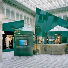 En marmorstaty i förgrunden på Medelhavsmuseet Stockholm. Gröna, geometriska installationer och antika föremål visas i den ljusa hallen.