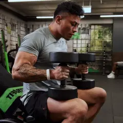 Mann hebt Hanteln für Muskelaufbau. Ein Trainer hilft ihm, während ein anderer Mann im Hintergrund trainiert.