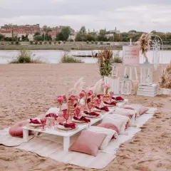 Elegancki wieczór panieński w plenerze nad rzeką. Stół z różowymi kieliszkami, serwetkami i kwiatami. W tle miasto.
