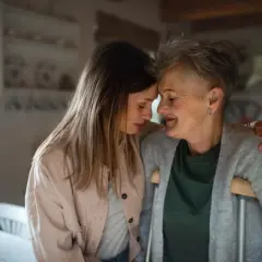 Dwie kobiety, matka i córka, dzielą się chwilą bliskości. Starsza kobieta, wspierana kulą, czule obejmuje młodszą. Obraz symbolizuje wsparcie w walce z chorobą Huntingtona.