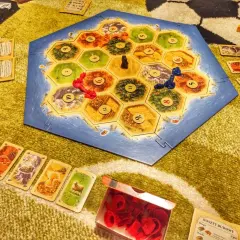 Plansza Catan: Świt Ludzkości, karty surowców i pionki. Rozgrywka w toku, gracze budują osady i drogi.