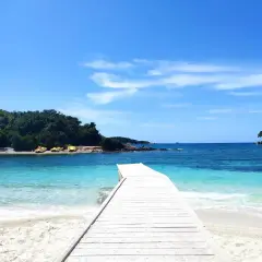 Drewniany pomost prowadzi do krystalicznie czystej wody na **Ksamil beach**. W oddali widać zielone wyspy i żółte łódki.