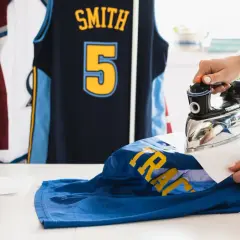 Une main utilise un fer à repasser pour défroisser un maillot de basket bleu avec des lettres jaunes. D'autres maillots sont suspendus en arrière-plan.