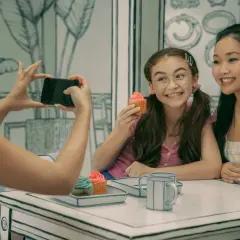 XO Kitty obsada: dwie dziewczynki jedzą babeczki, jedna robi zdjęcie telefonem.