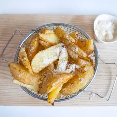 Cesta metálica llena de crujientes patatas gajo, espolvoreadas con queso rallado, sobre una tabla de madera.