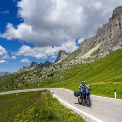 Motociclista percorre una strada panoramica tra i laghi del Veneto, con montagne imponenti e cielo azzurro.