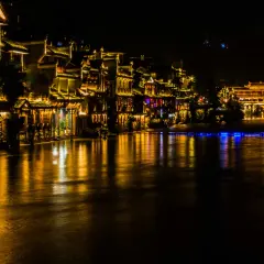 Nocny widok na Fenghuang: oświetlone tradycyjne budynki i most odbijające się w rzece.