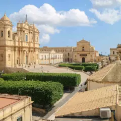 Piazza barocca con chiese imponenti e giardini curati, un esempio di città d'arte Italia sotto un cielo azzurro con nuvole bianche.