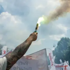 Dym z flary rozświetla niebo nad demonstracją związków zawodowych. W tle białe flagi z napisem "Solidarność".