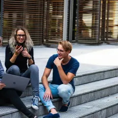 Drei junge Leute sitzen auf einer Treppe. Eine Frau mit Laptop und eine Freundin mit Handy, die sich über das Studium zuhause unterhalten.