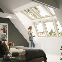 Jasny pokój na poddaszu z kobietą przy oknie. Dachowe okna wpuszczają dużo światła, tworząc przytulną atmosferę.