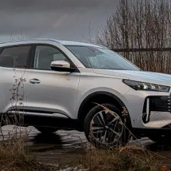 Srebrny Chery Tiggo 4 na tle ponurego krajobrazu. Auto stoi na mokrej nawierzchni, otoczone suchymi trawami i drzewami.