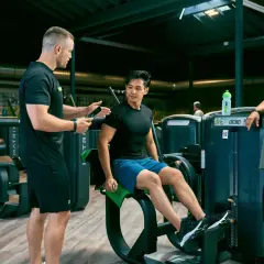 Trainer erklärt Mann die richtige Ausführung am Gerät für den erector spinae. Frau schaut zu.