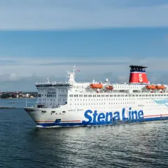 Statek Stena Line płynie po morzu, gotowy na rejs 24h. W tle widać wybrzeże z zabudowaniami.