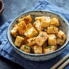 Usmażone kostki tofu, które jest zrobione z ziaren soi, w misce z sezamem i ziołami. Obok pałeczki i sos sojowy.