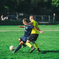 Dwóch piłkarzy walczy o piłkę na zielonej murawie. Jeden w żółtej koszulce próbuje odebrać ją przeciwnikowi, stosując agresywny pressing.