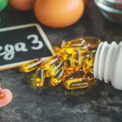 Kapsułki Omega-3 wysypujące się z białej butelki, na tle tabliczki z napisem "Omega 3".