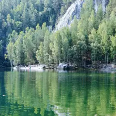 Zielone jezioro otoczone lasem i skalnymi formacjami. W oddali widać ludzi spacerujących po plaży. To malownicze miejsce, gdzie góry stołowe spotykają się z wodą.