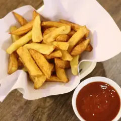 Chrupiące frytki stekowe w papierowym rożku obok miseczki z ketchupem na drewnianym tle.