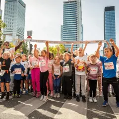 Dzieci z podniesionymi rękami, gotowe do startu w biegu. W tle wieżowce i hasła dopingujące biegaczy.