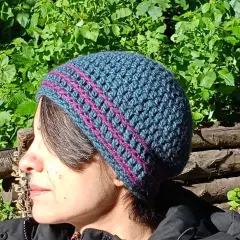 Donna con cappello uncinetto tutorial, blu e fucsia, con sfondo verde e tronchi.