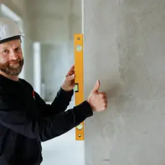 Budowlaniec w kasku sprawdza pionową ścianę za pomocą poziomicy. Kciuk w górę potwierdza, że wszystko jest idealnie.