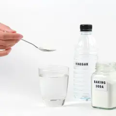 Une main tient une cuillère de bicarbonate de soude pour déboucher une canalisation. Vinaigre et eau sont prêts.