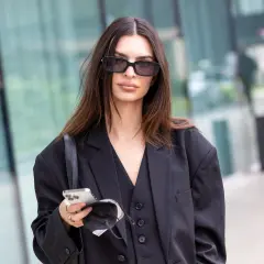 Elegancka brunetka w okularach przeciwsłonecznych, z telefonem i maseczką w dłoni. Jej włosy mają piękny, ciemny kolor.