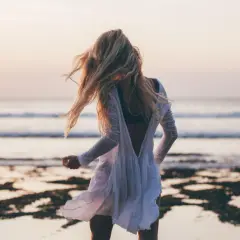 Blondynka w zwiewnej białej sukience tańczy na plaży, a wiatr rozwiewa jej włosy. W tle widać spokojne beach waves i zachodzące słońce.