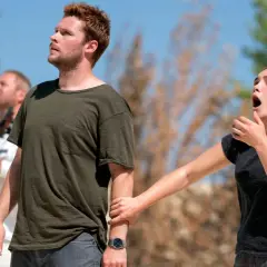 En kvinna griper tag i en mans arm, båda ser uppåt med förvånade uttryck. En man med ett instrument syns i bakgrunden. Scen från Midsommar filmens rollista.