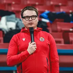 Właściciel Wisły Kraków w czerwonej bluzie z kapturem i mikrofonem, na tle pustych czerwonych trybun stadionu.