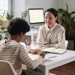 Ein Vater, sein Kind und eine freundliche Frau sitzen an einem Tisch. Die Frau, die wie eine Integrationshelferin aussieht, erklärt etwas. Das Kind malt. Das Gehalt eines Integrationshelfers ist eine wichtige Frage für viele.