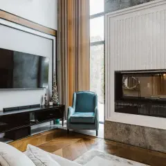 Nowoczesny salon z kominkiem, telewizorem i wygodnym fotelem. W tle widać okno z zasłonami, a na podłodze eleganckie spieki.