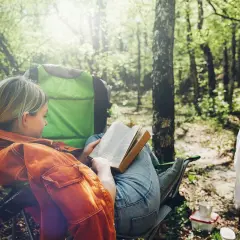 Dziewczyna czyta książkę w lesie, siedząc na krześle turystycznym. To idealne miejsce, by poznać zasady, jak zanocuj w lesie.