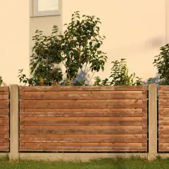 Drewniane deski tworzą ogrodzenie rustykalne, za którym widać zielone krzewy i fragment jasnego budynku.