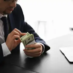 Mężczyzna w garniturze liczy banknoty dolarowe przy biurku. Jego praca w banku wymaga precyzji.